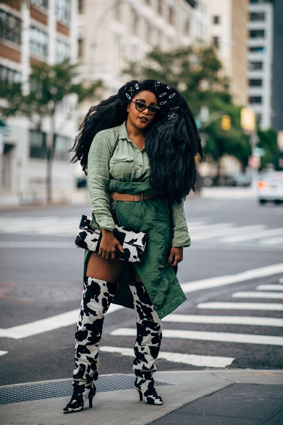 A curvy woman of color is rocking an outfit that features cow print boots with a perfectly coordinating clutch. Her clothing mixes various shades of green and accentuates her hourglass figure. She looks confident as she owns her Creative Style Archetype.